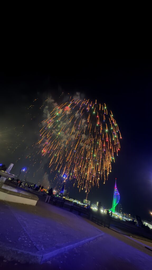 Colourful fireworks bursting in the night sky over a waterfront with people watching. John Rigby and Co