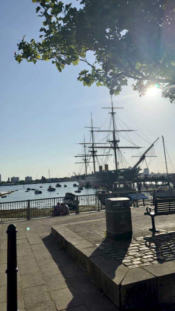 The Warrior at a dock with smaller boats and a clear blue sky, shadow of a tree in the foreground.