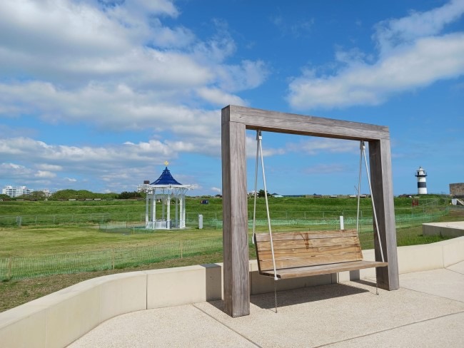 Swing bench, on a neutral paving stone with a grass background and a blue but lightly cloudy sky